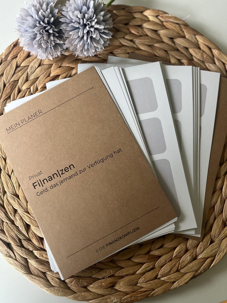K&ouml;nnte beinhalten: Ein braunes Notizbuch mit dem Text "MEIN PLANER" und "Privat Finanzen Geld, das jemand zur Verf&uuml;gung hat" auf dem Einband. Das Notizbuch liegt auf einer geflochtenen Weideunterlage mit zwei grauen Blumen.