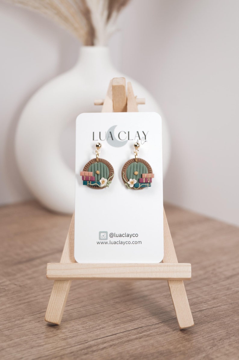 Op de afbeelding: Een paar groene deuroorbellen met een stapel boeken en bloemen. De oorbellen zijn gemaakt van polymer clay en hebben gouden haakjes. De oorbellen bevinden zich op een witte kaart met de tekst "LUA CLAY" en de social media handle "@luaclayco" en website "www.luaclayco.com".