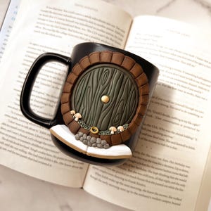 May include: Black ceramic mug featuring a detailed, three-dimensional design of a hobbit door. The door is green with a brown frame, a gold knob, and small mushroom accents. The mug is resting on an open book.