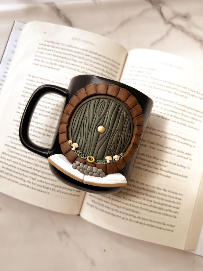 May include: Black ceramic mug featuring a detailed, three-dimensional design of a hobbit door. The door is green with a brown frame, a gold knob, and small mushroom accents. The mug is resting on an open book.