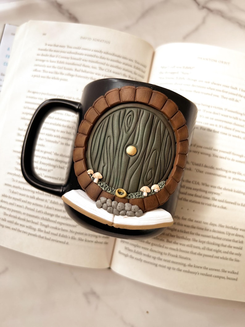 May include: A black ceramic mug featuring a detailed, handcrafted design of a hobbit door. The door is green with wood grain texture, surrounded by a brown frame. Small mushrooms and a ring are also part of the design. The mug rests on an open book.