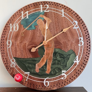 Puede incluir: Un reloj de madera con temática de golf. La esfera del reloj presenta a un golfista balanceando un palo, con acentos verdes y marrones. Los números son blancos y las manecillas del reloj son doradas. El reloj está diseñado para Farooq Ali.
