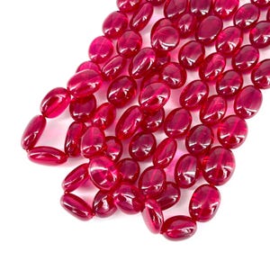May include: A collection of oval, translucent, deep red beads. The beads are smooth and polished, with a glossy finish. They are strung together, suggesting they are for jewellery making or crafting. The colour is consistent throughout, with a slight variation in tone.
