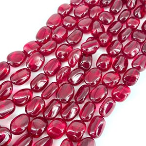 Puede incluir: Primer plano de una colección de cuentas pulidas, de forma ovalada, en un tono rojo intenso y translúcido. Las cuentas son lisas y reflectantes, lo que sugiere un material de vidrio o piedra preciosa, ideal para joyería.