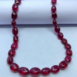 Puede incluir: Una hebra de cuentas de rubí de forma ovalada en un color rojo intenso. Las cuentas están enhebradas en un hilo transparente y están dispuestas en un patrón suelto y fluido.