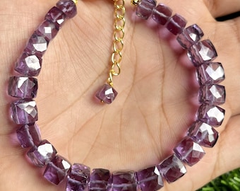 May include: A purple amethyst gemstone bracelet with a gold clasp and extender chain. The bracelet is made of faceted square beads.
