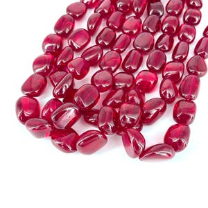 May include: A collection of polished, oval-shaped, deep red beads. The beads are translucent, with a glossy finish, and appear to be strung together. The beads are scattered across a white surface, creating a visually appealing display. Ideal for jewelry making.