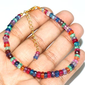 May include: A colorful beaded bracelet with a gold-colored clasp and extender chain. The bracelet features a variety of small, faceted beads in shades of red, pink, blue, green, and purple. The bracelet is displayed on a light-toned background.
