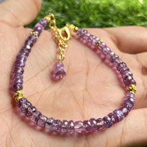 May include: A purple amethyst gemstone bracelet with a gold clasp and a small gold charm. The bracelet is made of small, faceted amethyst beads.