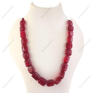 Puede incluir: Un collar de piedras preciosas rojas expuesto sobre un maniquí blanco. El collar está compuesto por cuentas cuadradas, cada una tallada con un diseño floral. Las cuentas están ensartadas, creando un bucle continuo. Las piedras preciosas tienen una calidad translúcida.