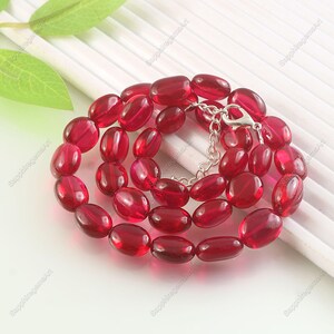 Puede incluir: Un collar con cuentas ovaladas translúcidas de color rojo intenso. Las cuentas están ensartadas en una cadena plateada con un cierre. El collar se exhibe sobre una superficie blanca, con una hoja en el fondo.