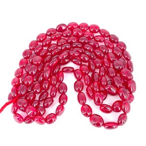 Puede incluir: Un conjunto de cuentas ovaladas de color rojo intenso. Las cuentas están ensartadas en un hilo rojo, formando una forma de collar. Las cuentas tienen un acabado pulido y brillante, que refleja la luz. El fondo es blanco liso.