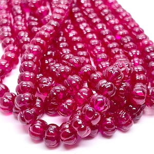 May include: A strand of bright pink, faceted, carved gemstone beads. The beads are round and have a textured, pumpkin-like design.
