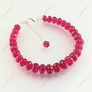 May include: A vibrant ruby red beaded bracelet with a silver clasp and extender chain. The beads are carved with a ribbed design. A small ruby flower charm hangs from the extender. Jewelry.