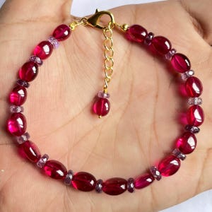 Puede incluir: Un brazalete de piedras preciosas rojas y moradas con un cierre y una cadena de extensión dorados. El brazalete está hecho de cuentas ovaladas.