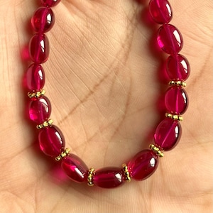 Pode incluir: Um bracelete de pedras preciosas vermelhas com detalhes dourados. O bracelete é feito de contas ovais e é enfiado em uma corrente dourada.