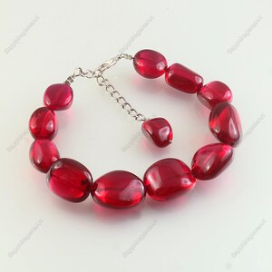 Puede incluir: Un brazalete de cuentas rojas con cuentas ovaladas. Las cuentas son de un rojo profundo y translúcido, unidas por una cadena y un cierre plateados. El brazalete se muestra sobre una superficie blanca.
