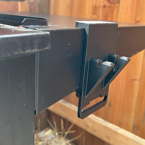 May include: Black metal bracket with two hooks, attached to a black metal surface.