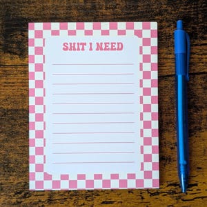 May include: A notepad with a pink and white chequered border and the text "SHIT I NEED" in pink. The notepad has lined pages and is accompanied by a blue pen.