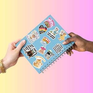 Pode incluir: Um caderno azul claro em espiral coberto por autocolantes coloridos. Os autocolantes apresentam v&aacute;rios desenhos, incluindo uma ch&aacute;vena de caf&eacute;, um gato, um OVNI e frases como "N&atilde;o fique triste!" e "Diz-me para sorrir mais uma vez."