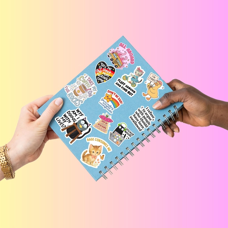 Pode incluir: Um caderno azul claro em espiral coberto por autocolantes coloridos. Os autocolantes apresentam v&aacute;rios desenhos, incluindo uma ch&aacute;vena de caf&eacute;, um gato, um OVNI e frases como "N&atilde;o fique triste!" e "Diz-me para sorrir mais uma vez."