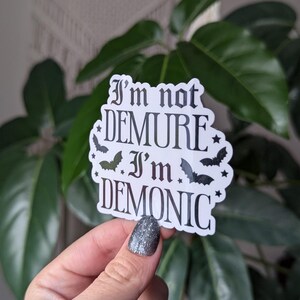 May include: A white sticker with black text that reads "I'm not demure I'm demonic" with bats and stars around the text.