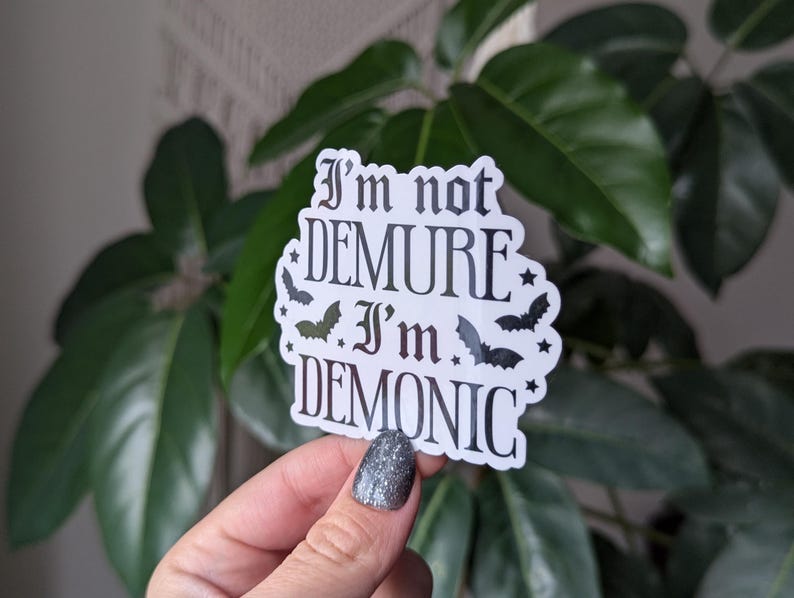 May include: A white sticker with black text that reads "I'm not demure I'm demonic" with bats and stars around the text.