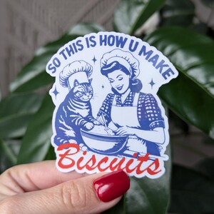 Puede incluir: Una pegatina azul y blanca con una ilustraci&oacute;n retro de una mujer y un gato haciendo galletas. El texto en la pegatina dice "So this is how you make biscuits".