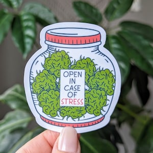 以下が含まれることがあります： 緑色のマリファナの花が詰まった瓶の漫画イラストが描かれた白いステッカー。瓶は赤い蓋と「ストレスの際は開けてください」と書かれた白いラベルが付いています。