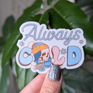 May include: A white sticker with blue and pink lettering that reads "Always Cold". A cartoon skeleton wearing a blue and orange hat and scarf is holding a blue cup with a white lid. The sticker has a white background with snowflakes.