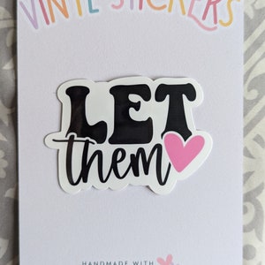 Op de afbeelding: Een witte vinyl sticker met de woorden "LET them" in het zwart, met een roze hart. De woorden zijn in een vetgedrukte, retro lettertype. De sticker zit op een witte kaart met de woorden "VINYL STICKERS" in pastelkleuren bovenaan.