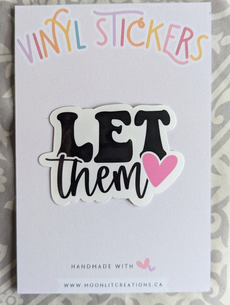 Op de afbeelding: Een witte vinyl sticker met de woorden "LET them" in het zwart, met een roze hart. De woorden zijn in een vetgedrukte, retro lettertype. De sticker zit op een witte kaart met de woorden "VINYL STICKERS" in pastelkleuren bovenaan.