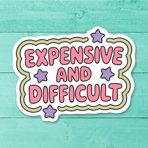 Peut inclure: Un autocollant blanc avec l'inscription "EXPENSIVE AND DIFFICULT" en lettres roses à bulles. Le texte est souligné en noir et entouré d'une bordure vert clair avec des étoiles violettes. Sur un fond en bois turquoise.