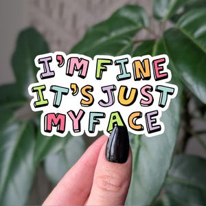 Op de afbeelding: Een kleurrijke sticker met de tekst "I'M FINE IT'S JUST MY FACE" in bubbelletters. De sticker heeft een witte rand en wordt voor een groene plant gehouden.