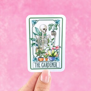 Puede incluir: Una pegatina blanca con un borde verde que muestra un esqueleto sosteniendo una planta en maceta. El esqueleto est&aacute; rodeado de otras plantas en maceta y flores. El texto "THE GARDENER" est&aacute; impreso en la parte inferior de la pegatina.