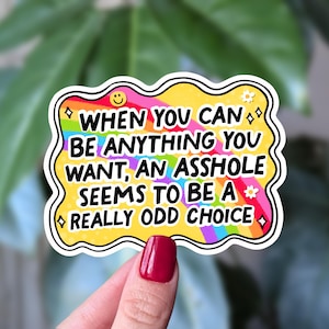 Peut inclure: Un autocollant coloré avec le texte "WHEN YOU CAN BE ANYTHING YOU WANT, AN ASSHOLE SEEMS TO BE A REALLY ODD CHOICE." L'autocollant a un fond jaune avec une bordure arc-en-ciel et un contour blanc.