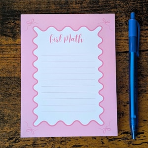 Op de afbeelding: Een roze notitieblok met een geschulpte rand en de woorden "Girl Math" in cursief. Een blauwe pen bevindt zich rechts van het notitieblok. Het notitieblok heeft horizontale lijnen om op te schrijven.