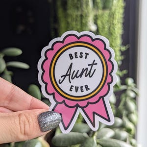 May include: A pink and yellow sticker with a ribbon. The sticker says "Best Aunt Ever" in a black and white font.