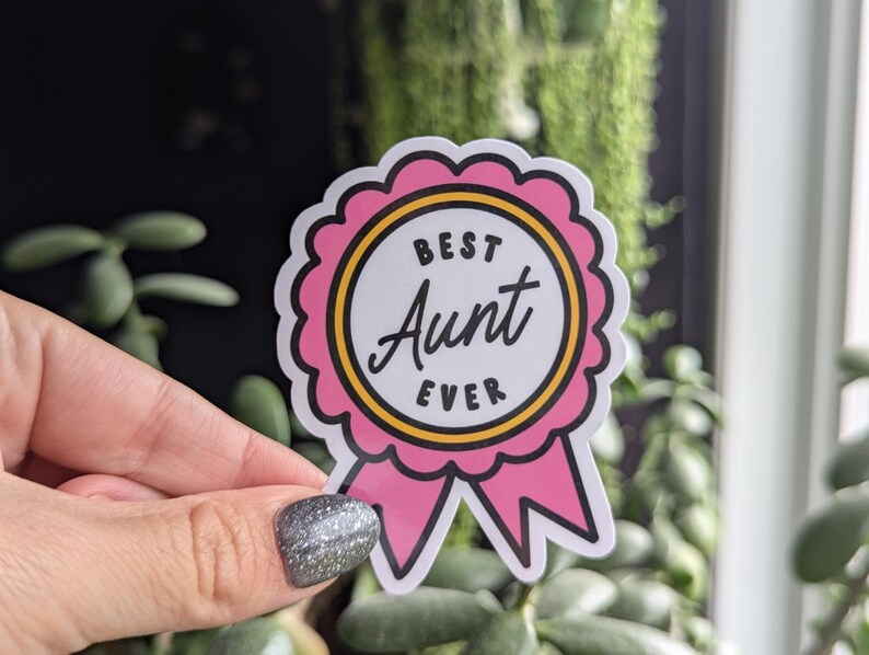 May include: A pink and yellow sticker with a ribbon. The sticker says "Best Aunt Ever" in a black and white font.