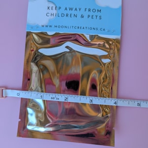 Może przedstawiać: Metaliczne, złote opakowanie z napisem "KEEP AWAY FROM CHILDREN & PETS" i adresem strony internetowej www.moonlitcreations.ca. Taśma miernicza pokazuje, że opakowanie ma szerokość około 10 cm.