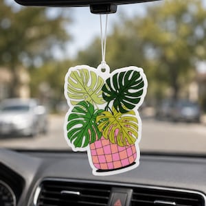 May include: Air freshener with a monstera plant design. The air freshener features a pink and white checkered pot with green and yellow leaves. It hangs from a white string, suspended from a car's rearview mirror.