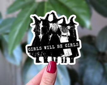 May include: A black and white sticker showing five figures in witch hats and dresses. The text "GIRLS WILL BE GIRLS" is on a white banner. The sticker is held by a hand with red nail polish.
