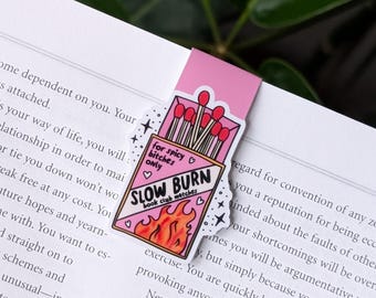 Könnte beinhalten: Ein pinkfarbener Lesezeichen mit einem Streichholzschachtel-Design. Auf der Streichholzschachtel steht "SLOW BURN book club matches" mit Flammen und dem Text "for spicy bitches only." Das Lesezeichen befindet sich auf einer Buchseite.