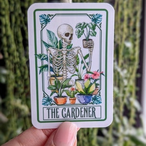 Puede incluir: Una pegatina blanca con borde verde muestra un esqueleto sosteniendo una planta en maceta. El esqueleto est&aacute; rodeado de otras plantas en maceta y flores. El texto "THE GARDENER" est&aacute; impreso en verde en la parte inferior de la pegatina.