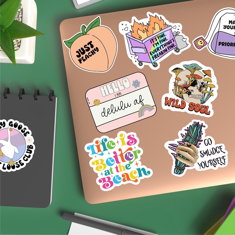 May include: A collection of colorful stickers featuring various designs, including a peach with the text "Just Peachy", a trash can with flames and the text "It's fine. I'm fine. Everything's freakin fine.", a skull with mushrooms and the text "Wild Soul", a hand holding a bundle of sage with the text "Go Smudge Yourself", and a rainbow text design that says "Life is Better at the Beach".