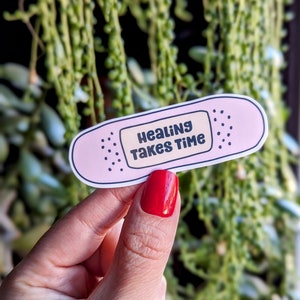 Pu&ograve; includere: Etichetta adesiva a forma di cerotto rosa con il testo "Healing Takes Time" in lettere nere.