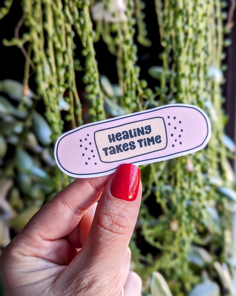 Pu&ograve; includere: Etichetta adesiva a forma di cerotto rosa con il testo "Healing Takes Time" in lettere nere.