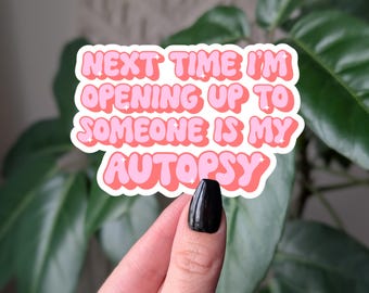 May include: A sticker with the text "NEXT TIME I'M OPENING UP TO SOMEONE IS MY AUTOPSY" in pink bubble letters with a white outline. The sticker is held by a hand with a black painted nail, against a background of green leaves.