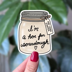 May include: A die-cut sticker shaped like a glass mason jar filled with beige sourdough starter. The jar features the black text 'I'm a hoe for sourdough' with a small heart and a tag reading 'HOE-MADE STARTER' tied with a string around the lid.