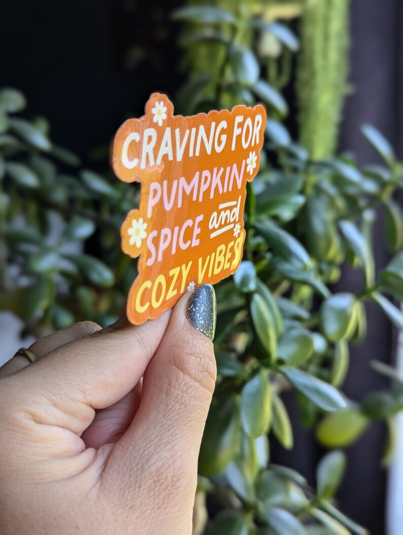 May include: Orange sticker with white text that reads "Craving for Pumpkin Spice and Cozy Vibes". The sticker has a daisy design around the text.